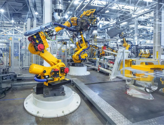 Industrial Robots With Yellow Arms Work On An Assembly Line In A High-Tech Manufacturing Factory. The Environment Is Clean And Well-Lit, With Various Machinery And Equipment Visible In The Background. The Robots Are Engaged In Automated Production Tasks.