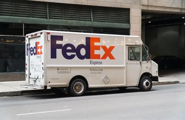 A White Fedex Delivery Truck Is Parked On The Side Of An Urban Street. The Truck Has The Fedex Logo Prominently Displayed On Its Side In Purple And Orange Letters. The Background Includes A Sidewalk, Buildings, And A Partially Closed Garage Door.