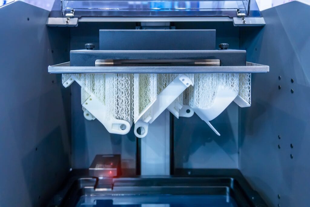 A 3D Printer Creates Intricate Parts With White Filament. The Partially Completed Pieces Hang Upside Down, Featuring Fine Mesh And Loop Designs. The Interior Of The Printer Is Visible, Showcasing Its High-Tech Components.