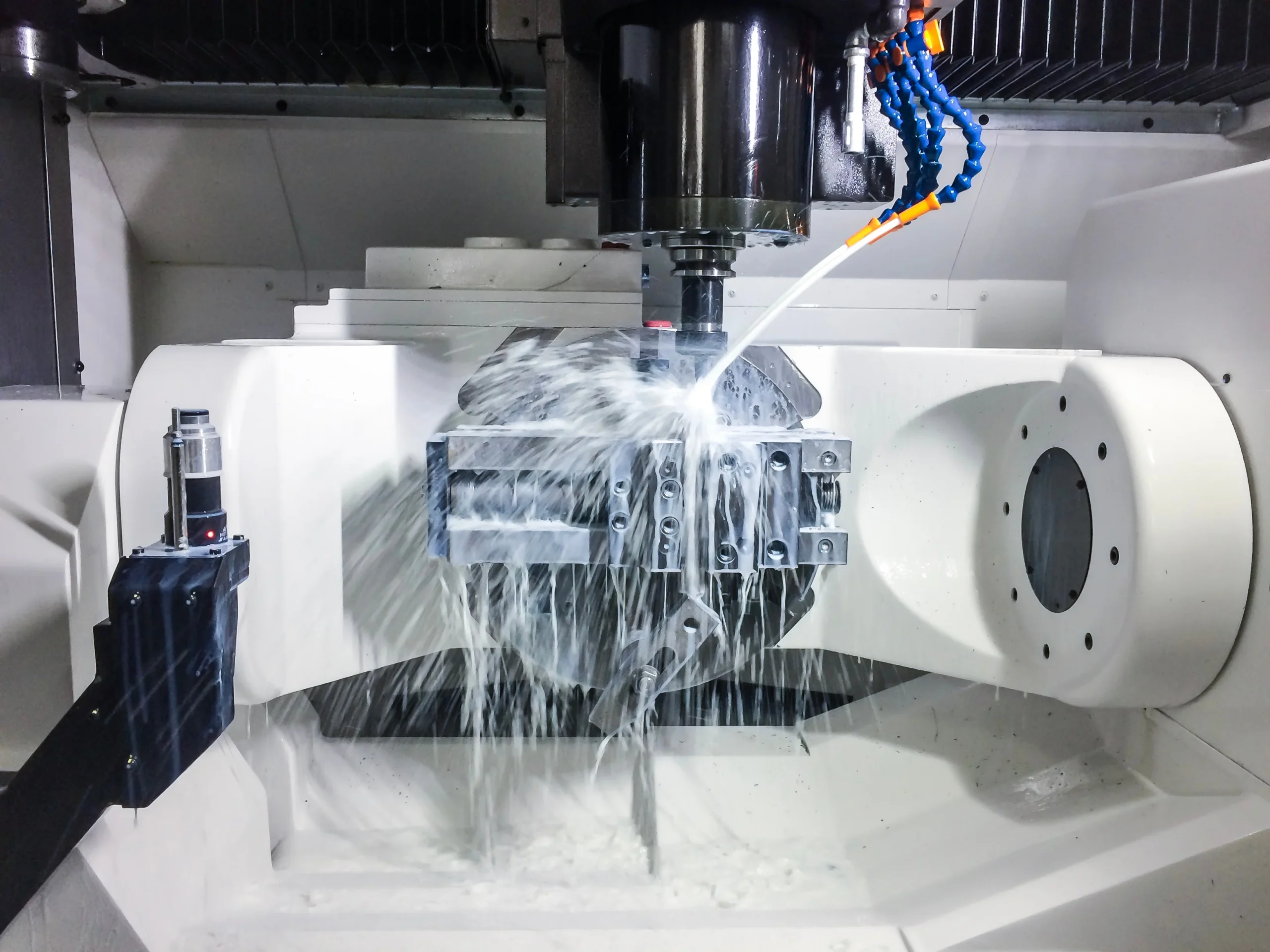 A CNC milling machine in operation, spraying coolant over a metal workpiece being machined. The machine has a rotating spindle and a fixture holding the workpiece, all mounted within a protective enclosure.