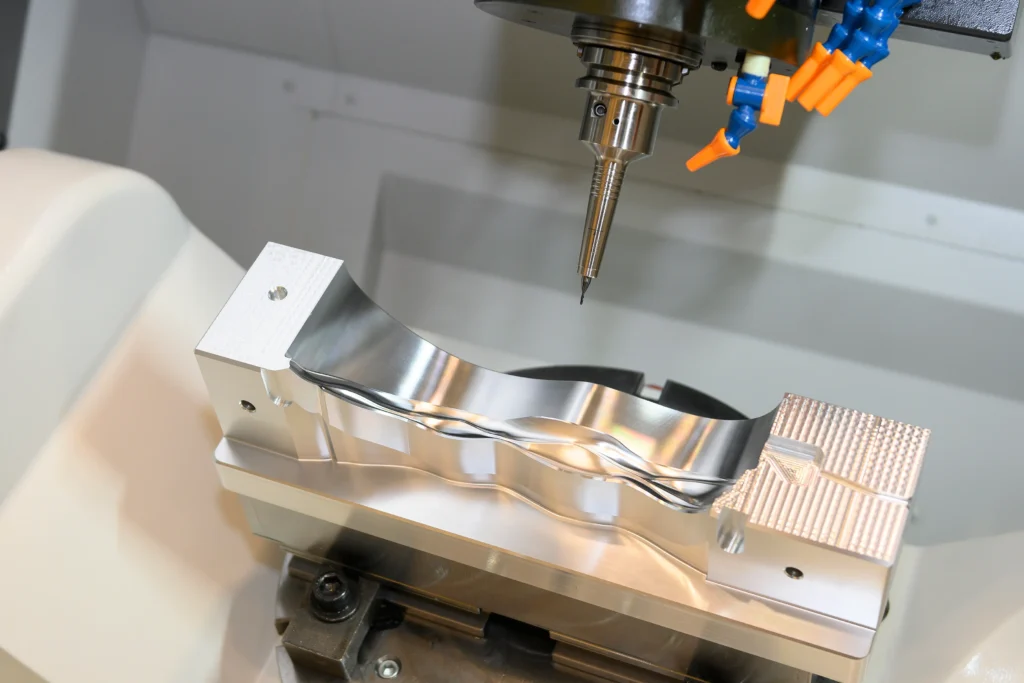 Close-Up Of A Cnc Machine Milling A Shiny Metal Component. The Device's Drill Bit Is Positioned Above The Workpiece, Showcasing Precision Engineering. Bright Metal And Sharp Tooling Feature Prominently In The Image.