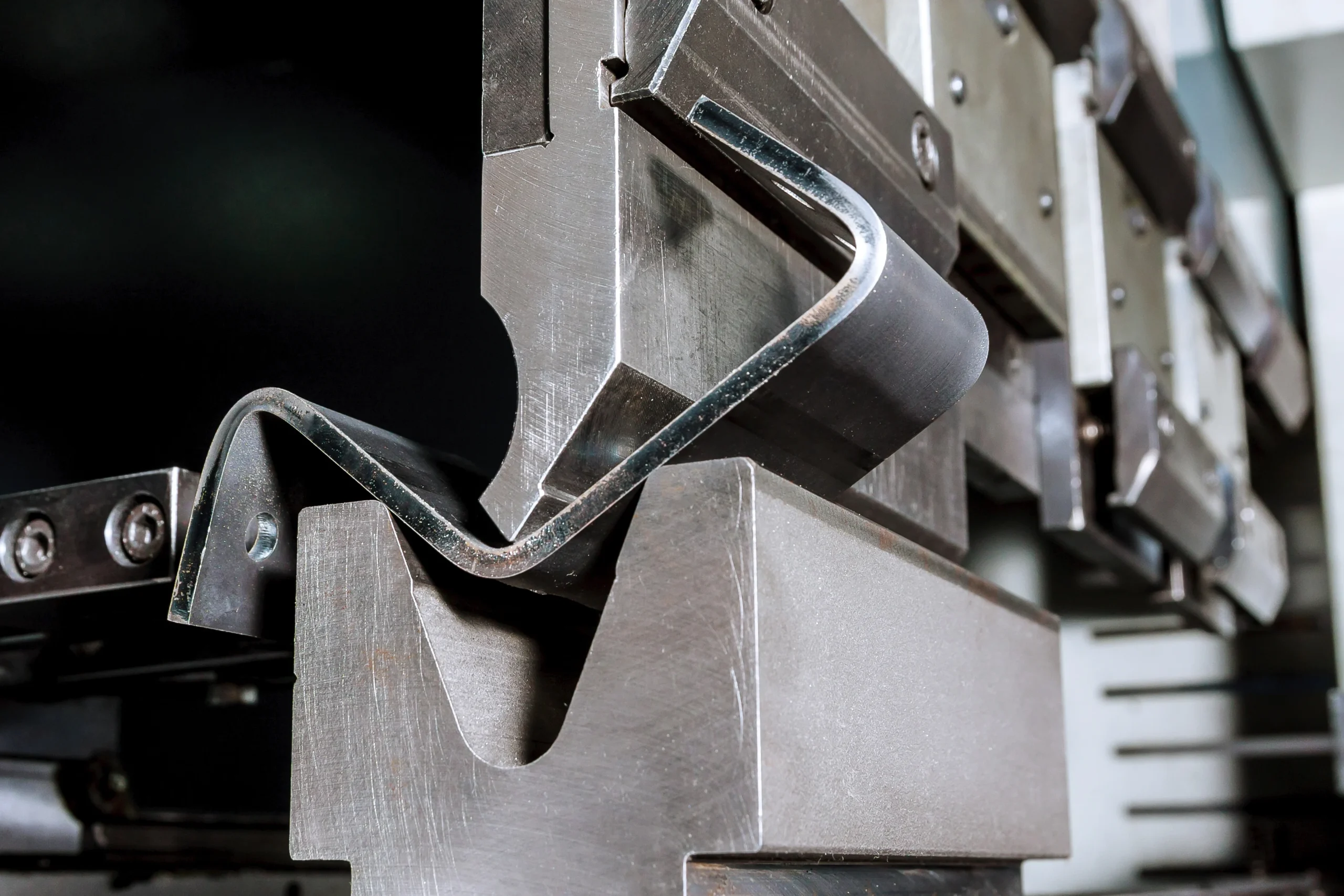 Close-up of a metal press brake machine bending a sheet of metal into a precise angle. The metallic surfaces are shiny, and the intricate details of the machine's components are visible, illustrating the industrial process.