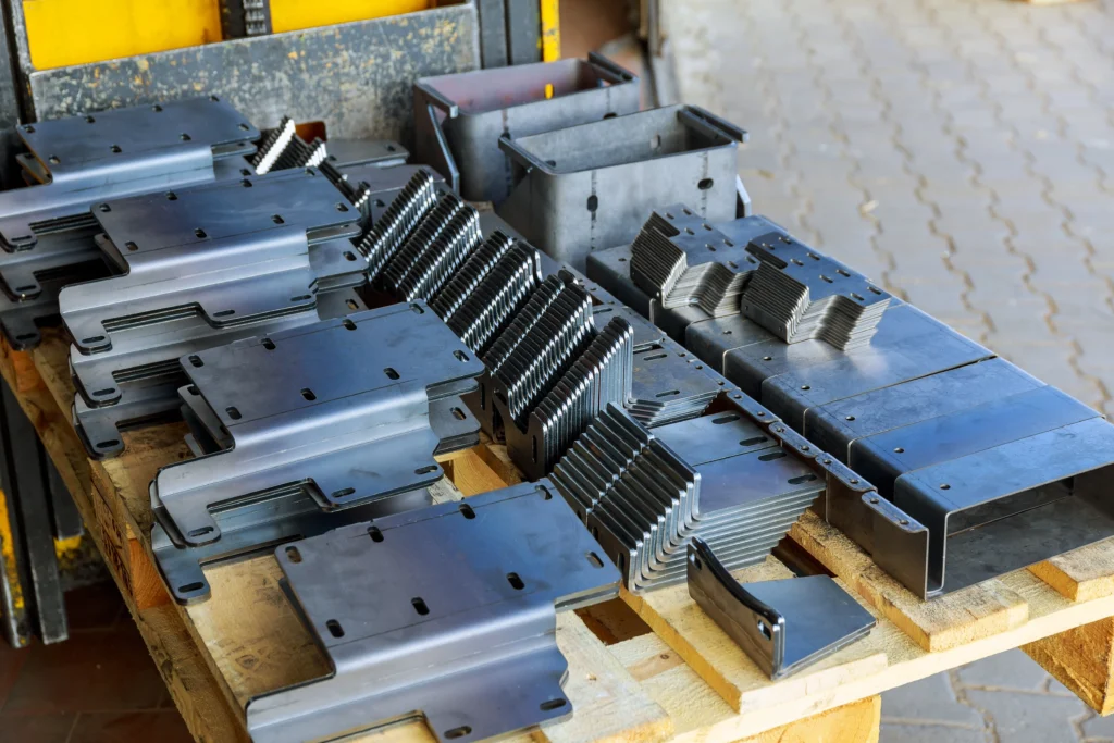 A Variety Of Metal Parts And Components Neatly Arranged On A Wooden Pallet. The Pieces Display Different Shapes And Sizes, Including Angled Brackets And Flat Panels, Showcasing Precision Machining. The Background Features Paving Stones.