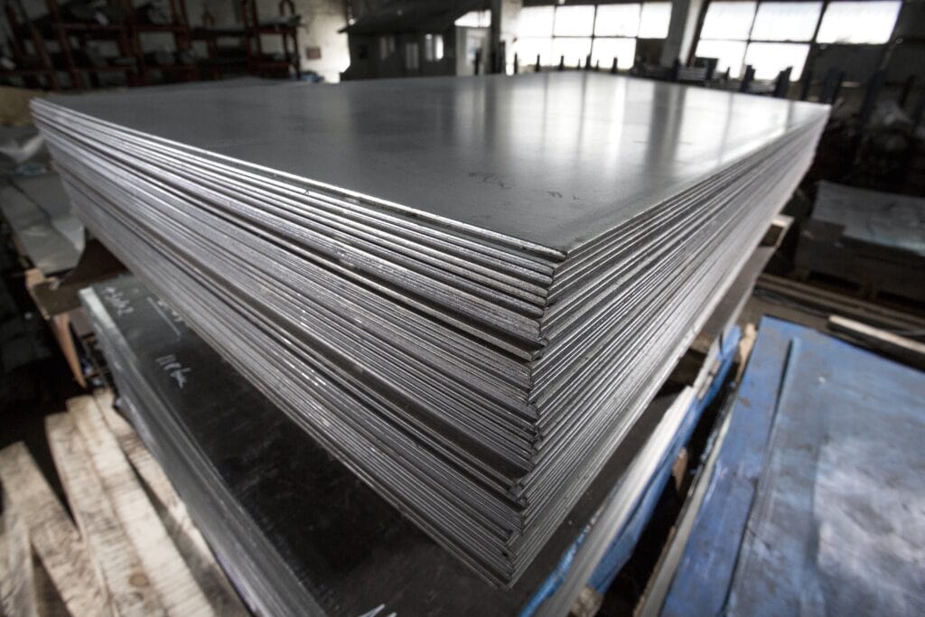 A Stack Of Large, Shiny Metal Sheets Is Shown In A Warehouse. The Sheets Are Neatly Piled On A Wooden Pallet, With Industrial Metal Racks And Windows Visible In The Background.