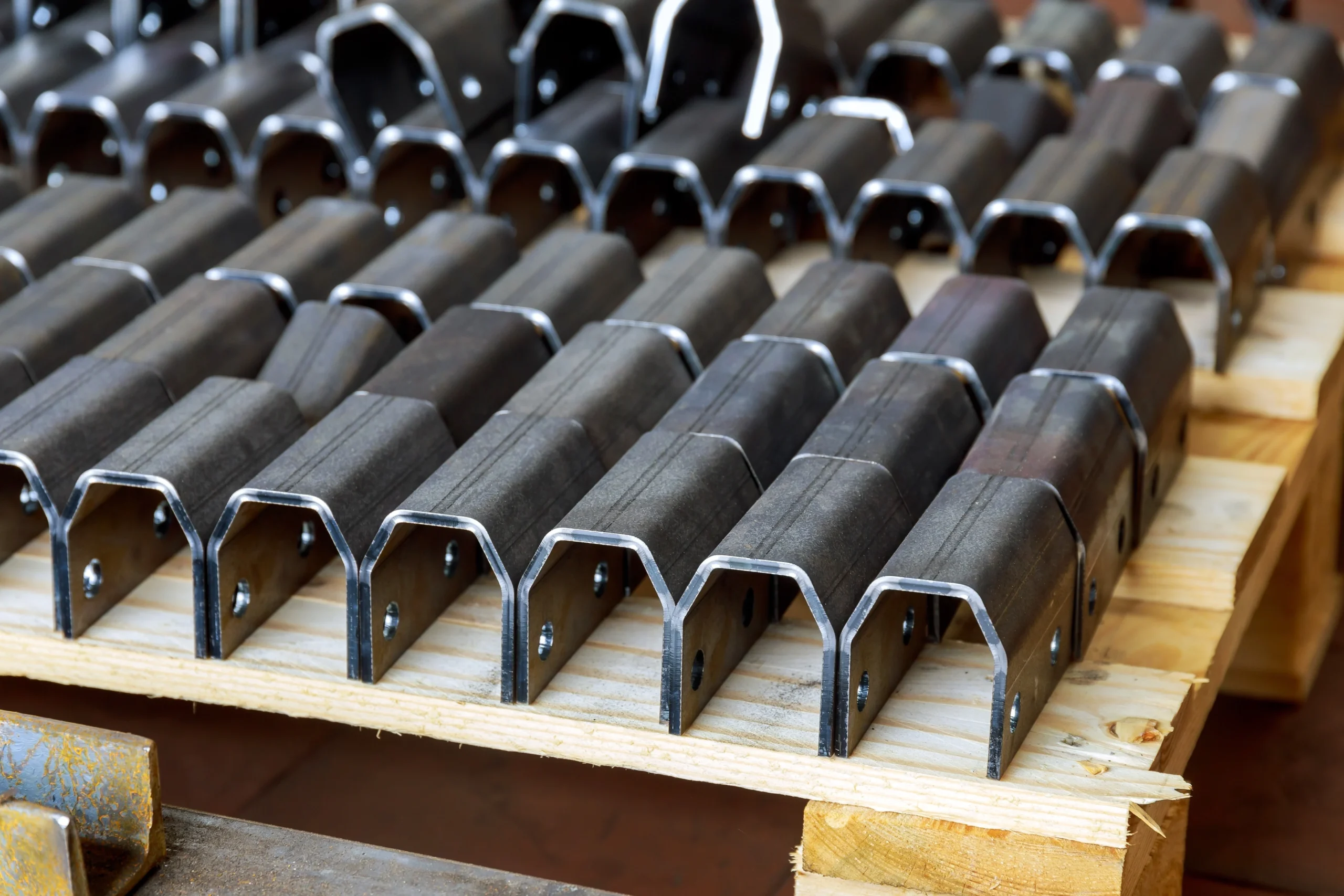 A close-up of neatly arranged metal brackets on a wooden pallet. The brackets are aligned in rows, each with a U-shape and small holes for screws or bolts. The wooden pallet provides a sturdy base for the shiny, industrial metal pieces.