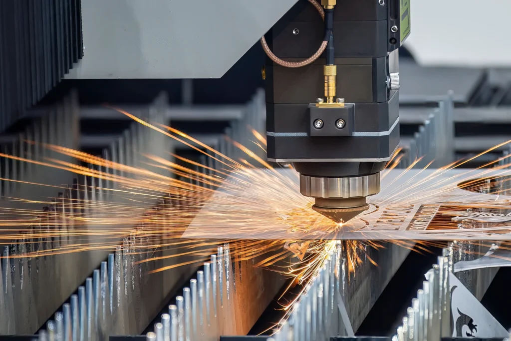 A Close-Up Of A Laser Cutting Machine In Action, Emitting Bright Sparks As It Cuts Through A Sheet Of Metal. The Focused Laser Beam Is Surrounded By Machinery Parts On A Metalwork Surface.