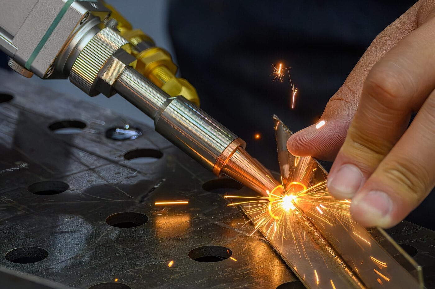 Close-up of a person's hands using a welding torch on a metal surface, with bright sparks flying. The metal piece is placed on a perforated metal table. The torch is pointed at the intersection of two metal pieces.