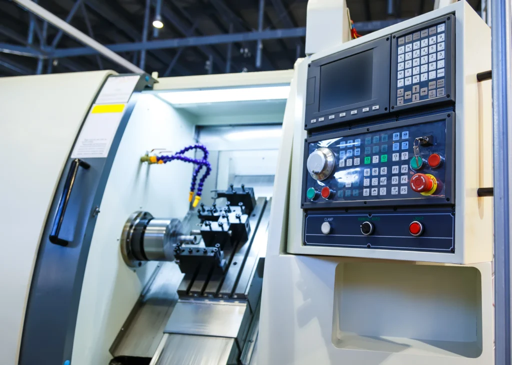 A Cnc Machine With An Open Compartment Displaying A Metal Component Being Worked On. The Control Panel To The Right Has Numerous Buttons And A Display Screen, Situated In An Industrial Setting With Overhead Lighting.