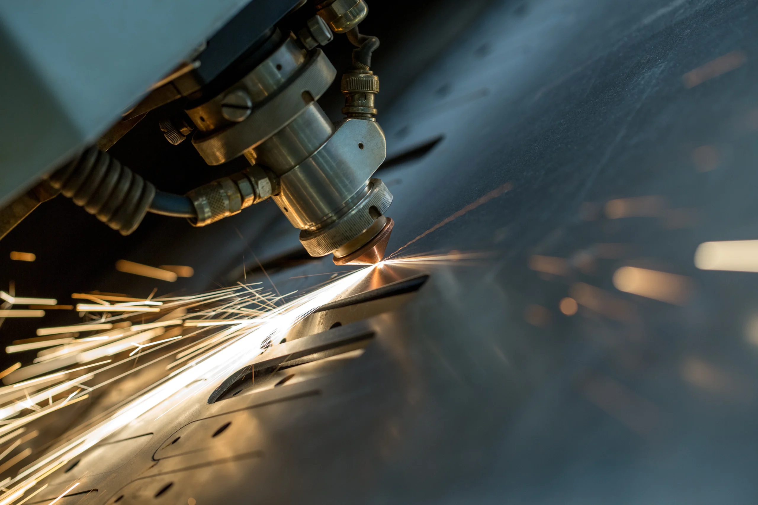 Close-up of a laser cutting machine in action, emitting bright sparks as it cuts through a sheet of metal. The metallic surface reflects the light of the sparks, highlighting precision engineering.