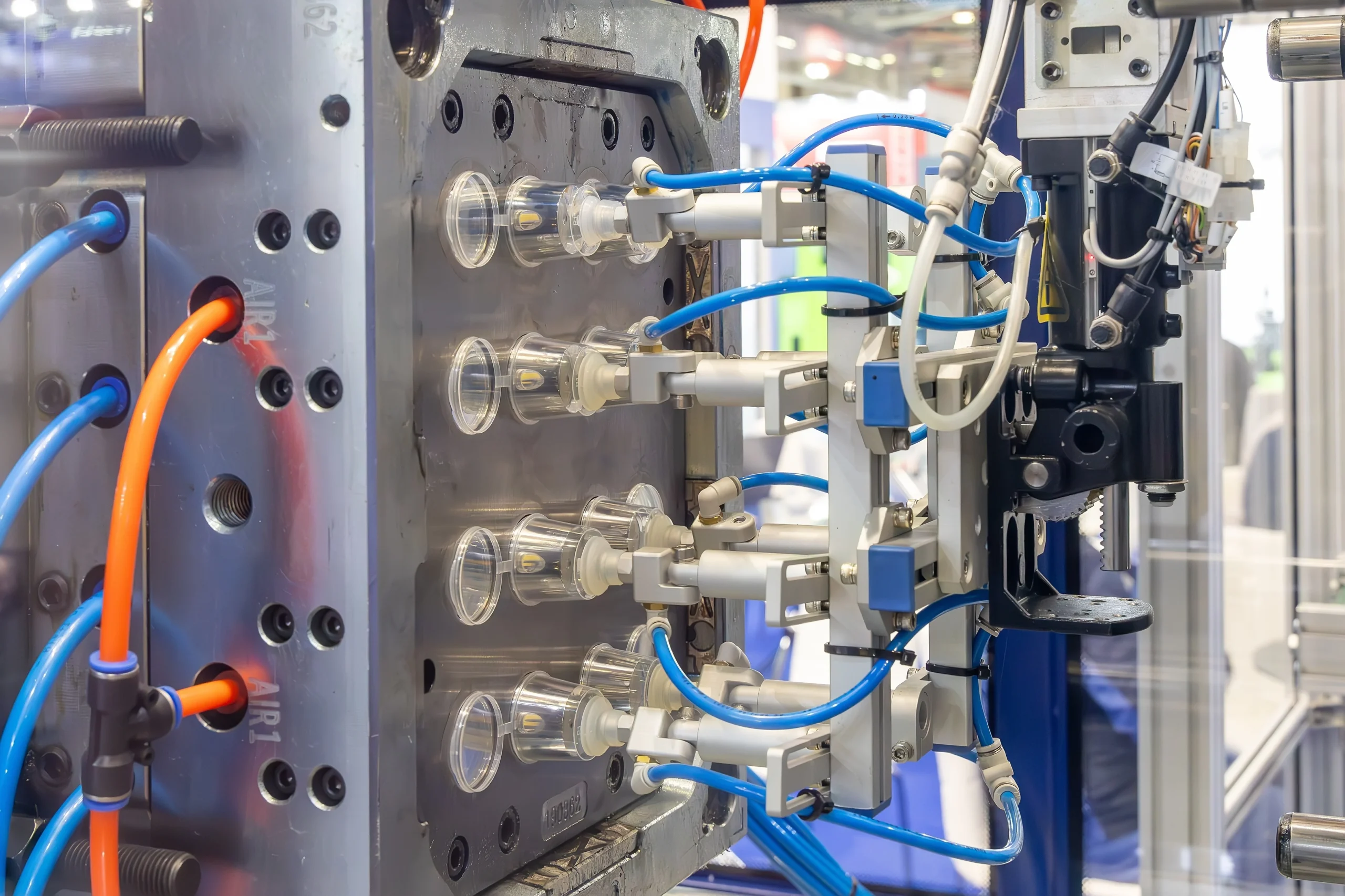 Close-up of an industrial machine used for plastic injection molding. The image shows several molds and connected tubes in various colors, including blue and orange. The setting appears to be a factory or manufacturing facility.