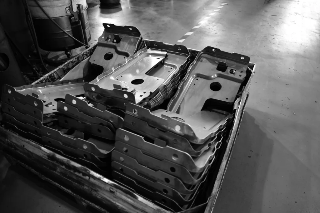 A Stack Of Metal Parts Lies In A Container On An Industrial Floor. The Parts Are Uniformly Shaped With Several Holes And Angles, Ready For Assembly Or Processing. The Setting Suggests A Manufacturing Or Mechanical Environment. Black And White Image.