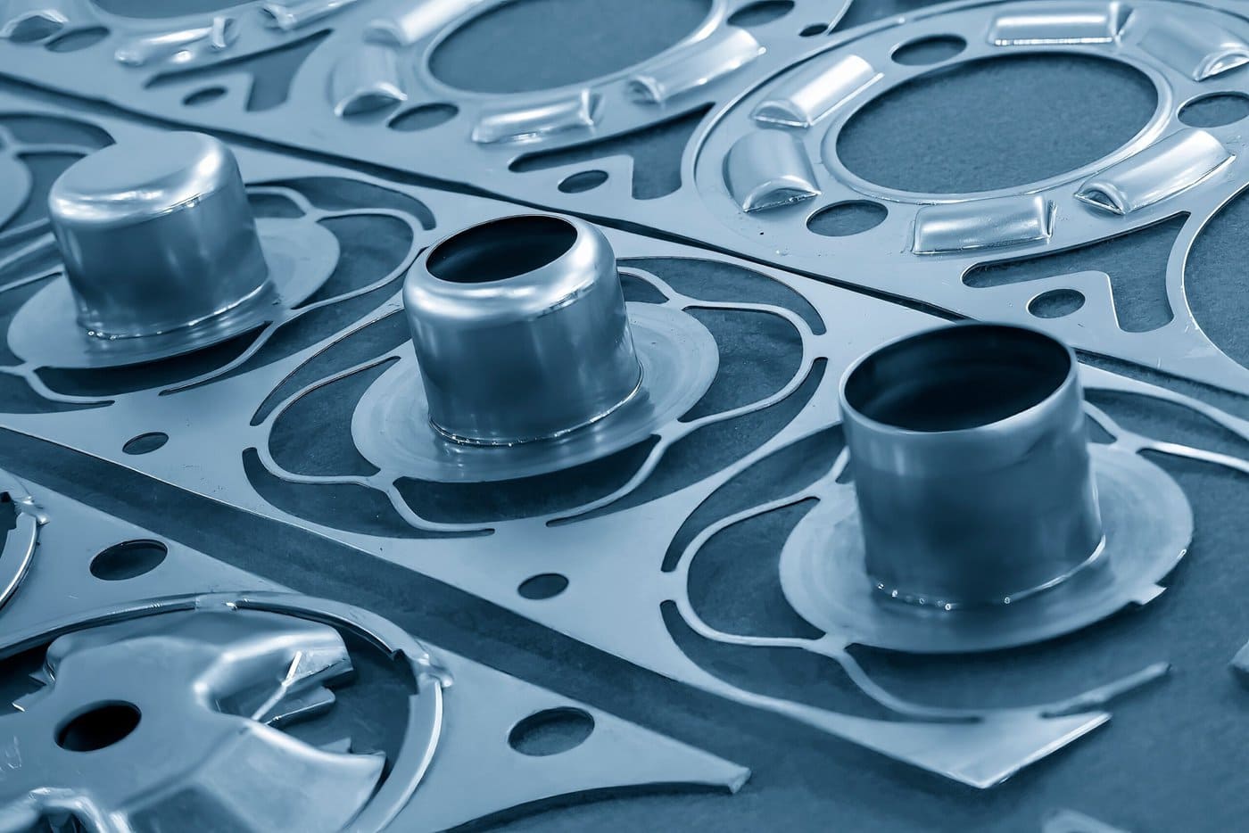 Close-up of precision-engineered metal components arranged in a symmetrical pattern. The parts feature circular cutouts and cylindrical protrusions, displaying a metallic sheen and intricate design against a flat, neutral background.