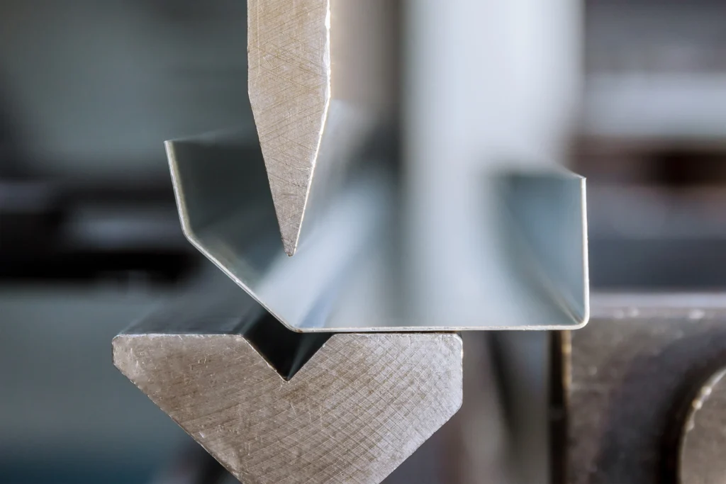 Close-Up Of A Sheet Metal Bending Process, Showing A V-Shaped Die And A Punch Pressing Down To Form A Precise Angle. The Metal Is In Focus, Highlighting The Detail Of The Bending Machinery And The Smooth Finish Of The Metal.