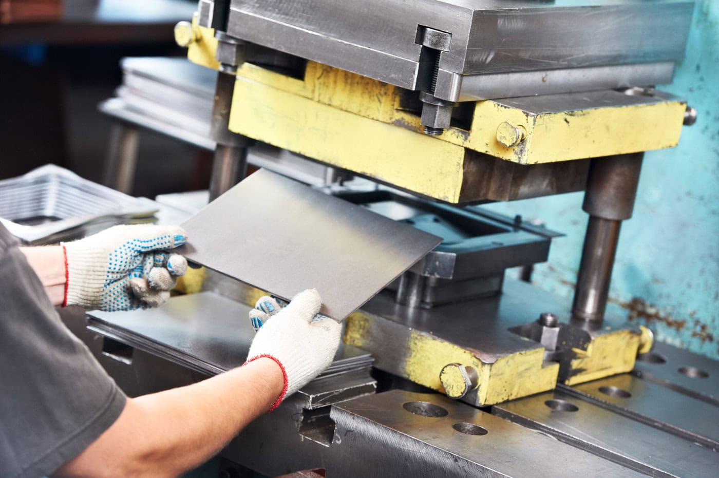 A worker wearing gloves operates a metal stamping machine, carefully placing a sheet of metal into the press in an industrial setting. The machine has yellow and gray components.