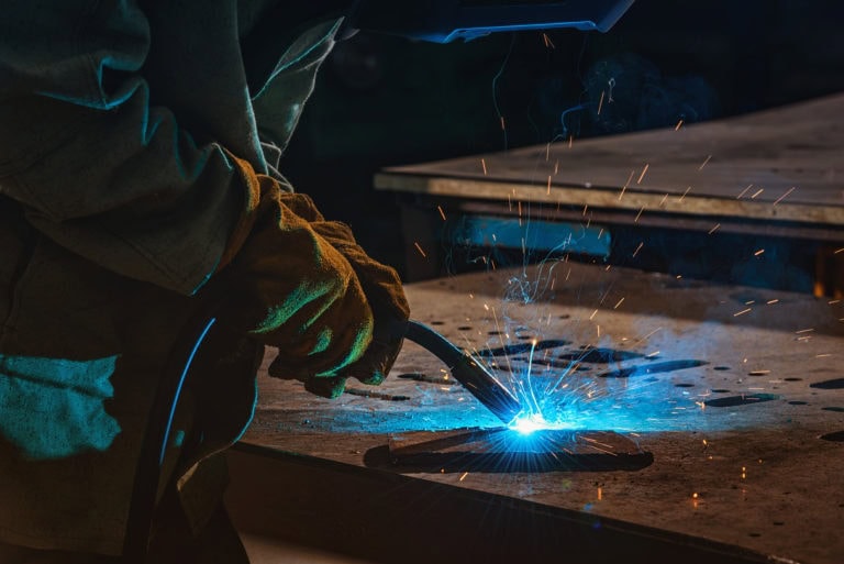 A person wearing protective gloves and a helmet is welding metal in a dimly lit workshop. Bright blue sparks and light emanate from the welding torch, highlighting the metal surface and creating an intense contrast with the dark background.