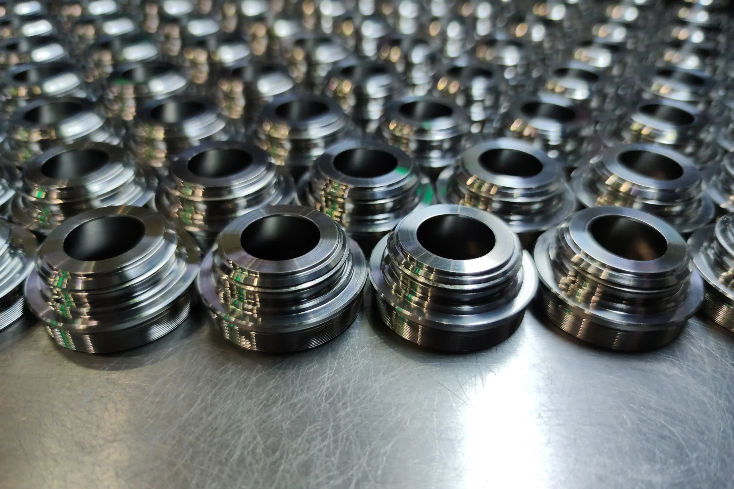 Close-up of rows of shiny metal components with multiple circular layers, arranged neatly on a reflective surface. The lighting highlights their polished texture, creating a visually intriguing pattern.