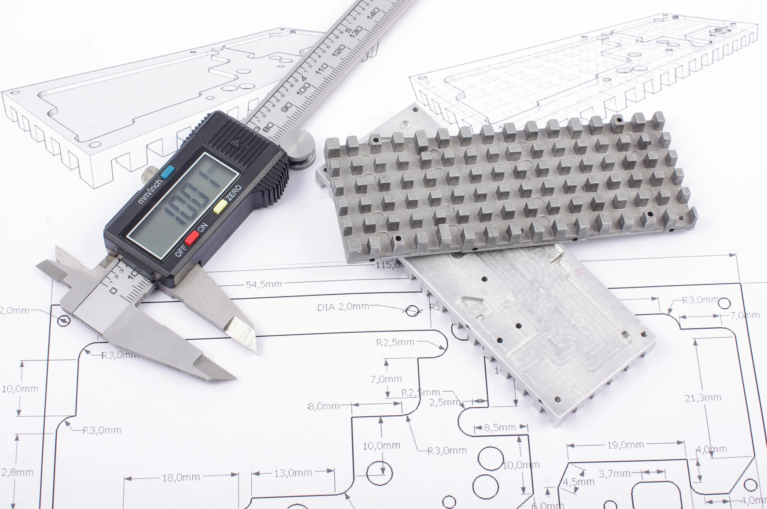 A caliper and metal components lie atop technical blueprints. The caliper's digital screen displays a measurement, and the blueprints feature detailed diagrams with dimensions, suggesting an engineering or manufacturing project.