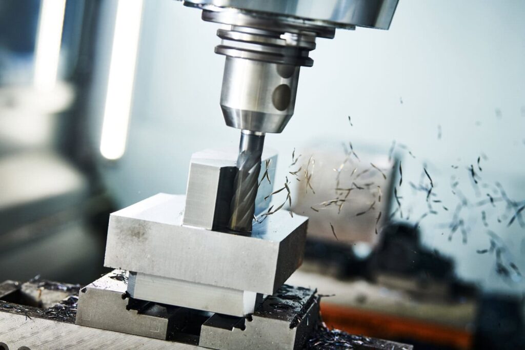 A Metal Milling Machine In Action, Cutting Into A Block Of Metal. Metal Shavings Fly Off As The Drill Bit Rotates. The Surrounding Area Is Slightly Blurred, Emphasizing The Motion And Precision Of The Machining Process.