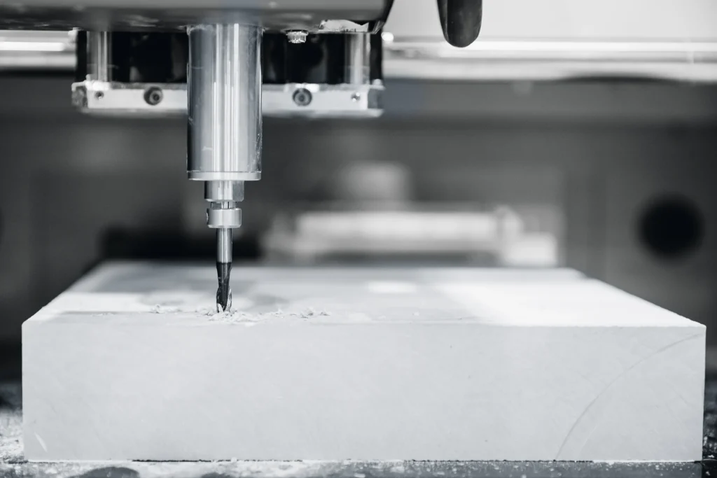 Close-Up Of A Cnc Machine In Operation, Carving Into A Rectangular Block Of Material. The Drill Bit Is Actively Engaged, Producing Visible Shavings Around The Area Being Cut. The Image Is In Black And White, Emphasizing The Machine'S Precision.