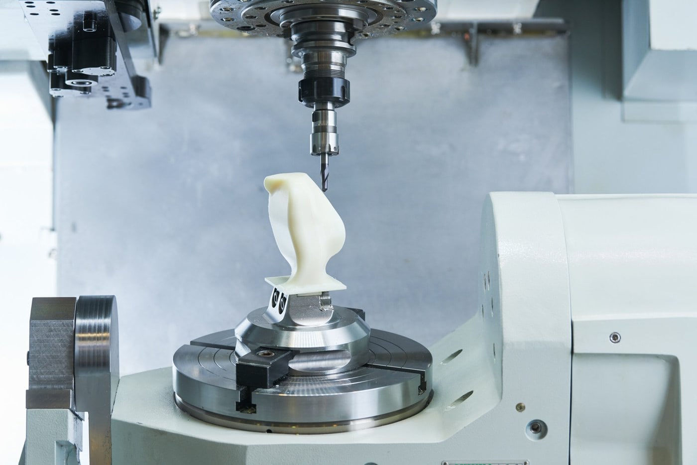 A CNC machine is precisely carving a white 3D object on a rotating platform. The machine's tool is in motion above the object, focusing on intricate details. The setup is inside a well-lit, industrial space.