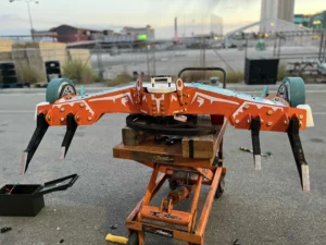 A Large, Angular Robot With Orange And Black Coloring Sits On An Orange Cart Outdoors. The Robot Features Long, Pointed Appendages And Wheels, Set Against An Industrial Backdrop With Buildings In The Distance.