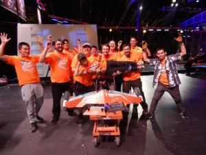 A Group Of People In Orange Shirts Are Joyfully Posing Around A Robot. One Person Is Being Held Horizontally Across The Group. They Are On A Stage With An Industrial And Tech-Like Setting.