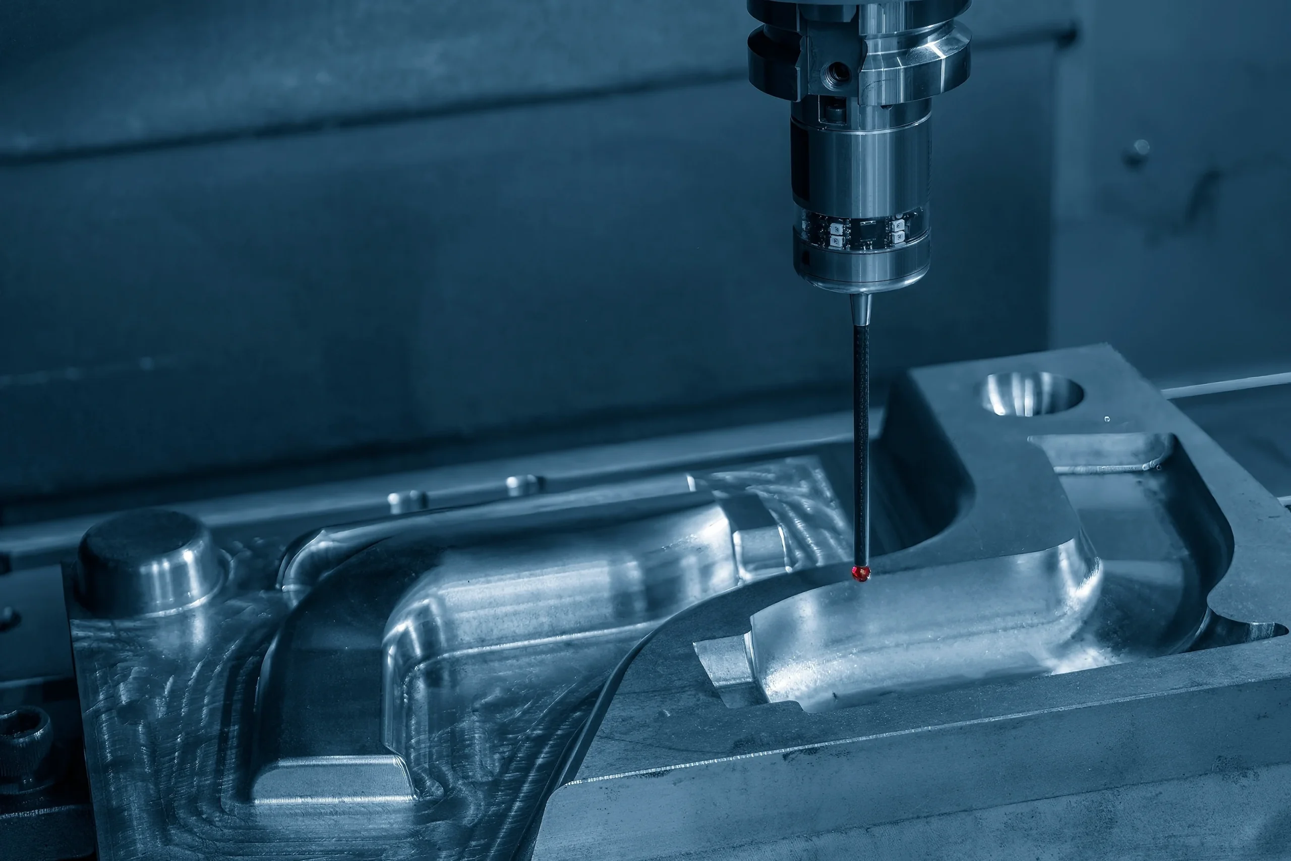 A CNC machine with a precision drill is cutting a detailed metal part. The setup is illuminated with a blue tint, highlighting the smooth, shiny surface of the metallic piece. The environment suggests a high-tech manufacturing process.