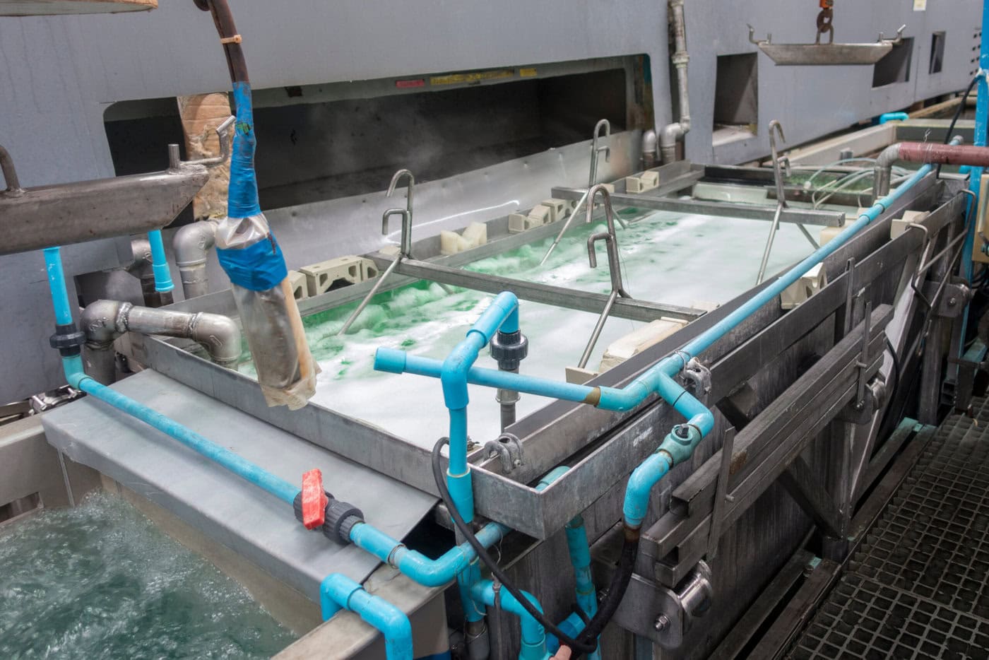 Industrial equipment with blue pipes and tanks processing green and white liquid. The setup appears to be part of a manufacturing or treatment plant, with various connectors and valves visible.