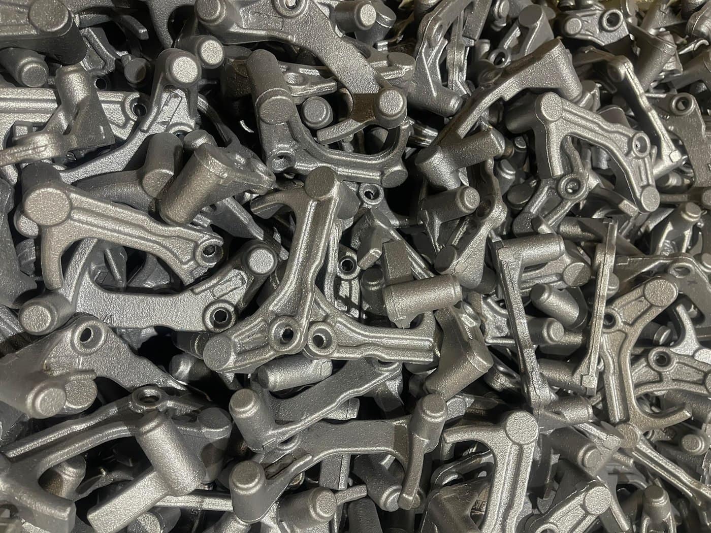 A pile of metal industrial parts with a matte gray finish, featuring various geometric shapes and holes, stacked closely together. The parts have a textured surface and appear freshly manufactured.