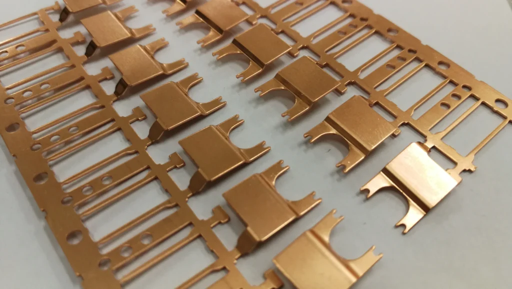 Close-Up Of A Series Of Copper-Colored Metal Components Aligned In Rows On A Thin Perforated Sheet, Likely Part Of A Manufacturing Or Assembly Process. The Components Have A Flat, Rectangular Shape With Prongs On One Side.