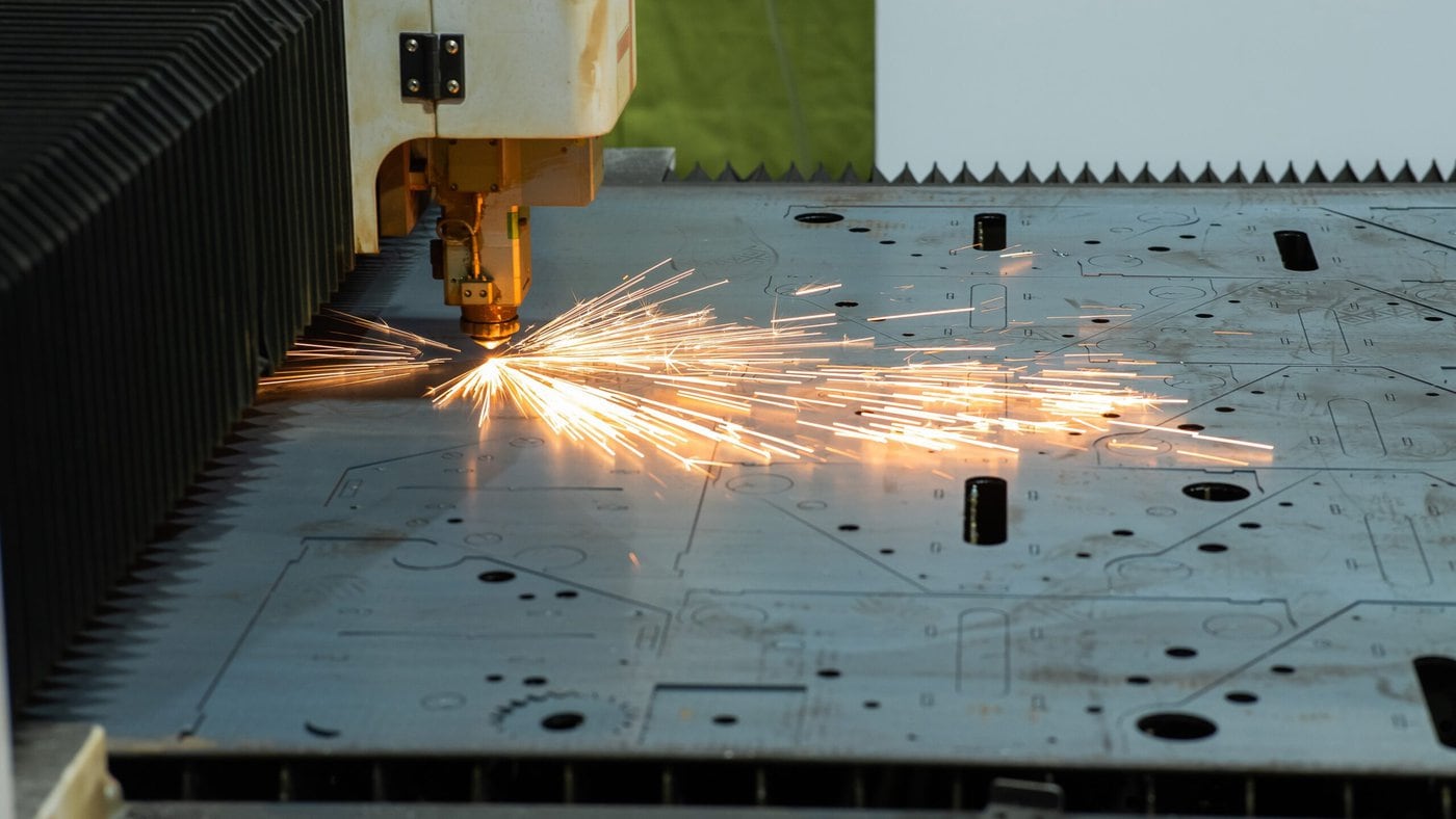 A laser cutting machine processing a sheet of metal, producing bright sparks. The metal surface is partially cut with visible patterns and holes, while the machine's nozzle is in action.