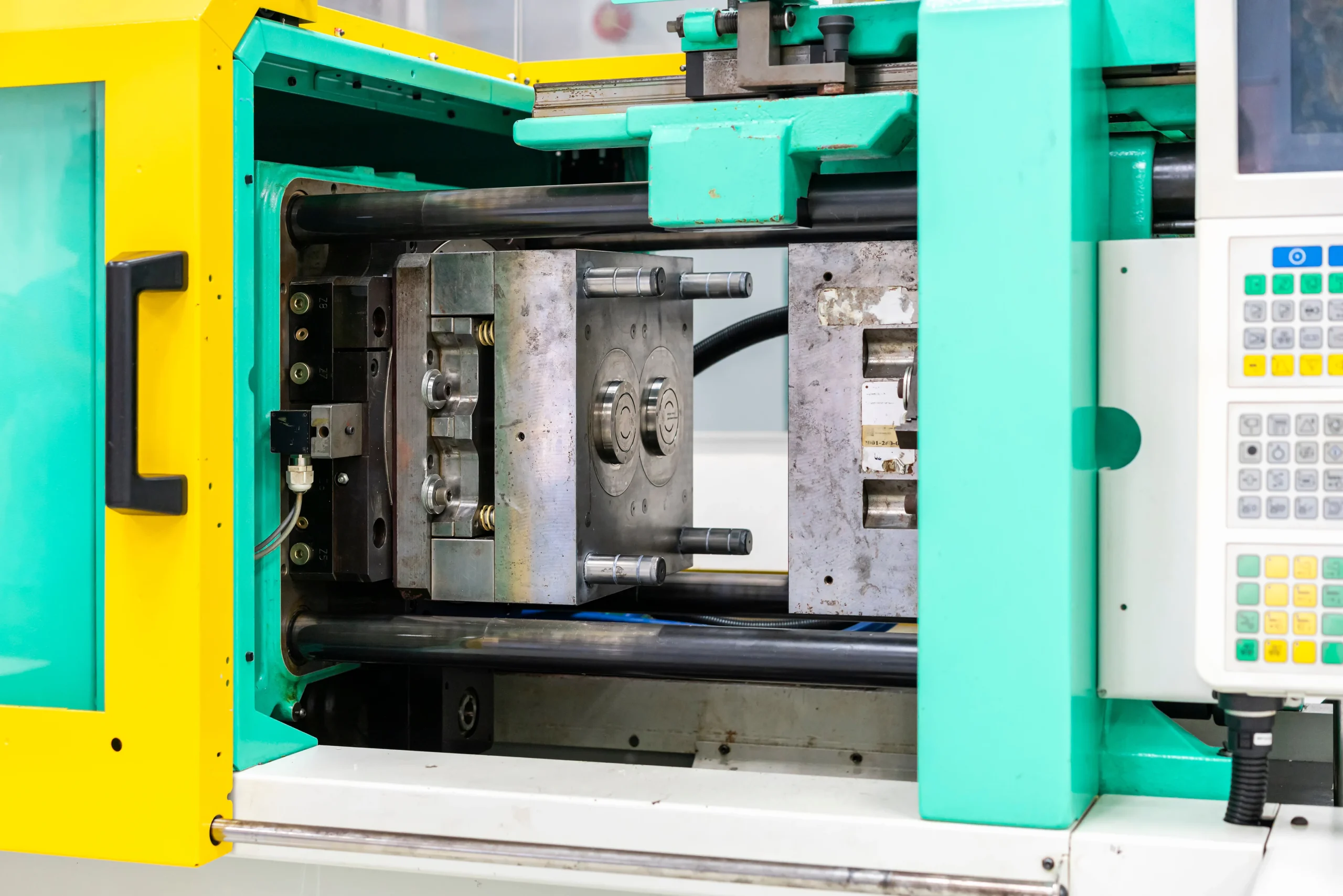 Close-up of an injection molding machine with an open mold. The machine has bright green and yellow panels and metal components, with a control panel visible on the side.