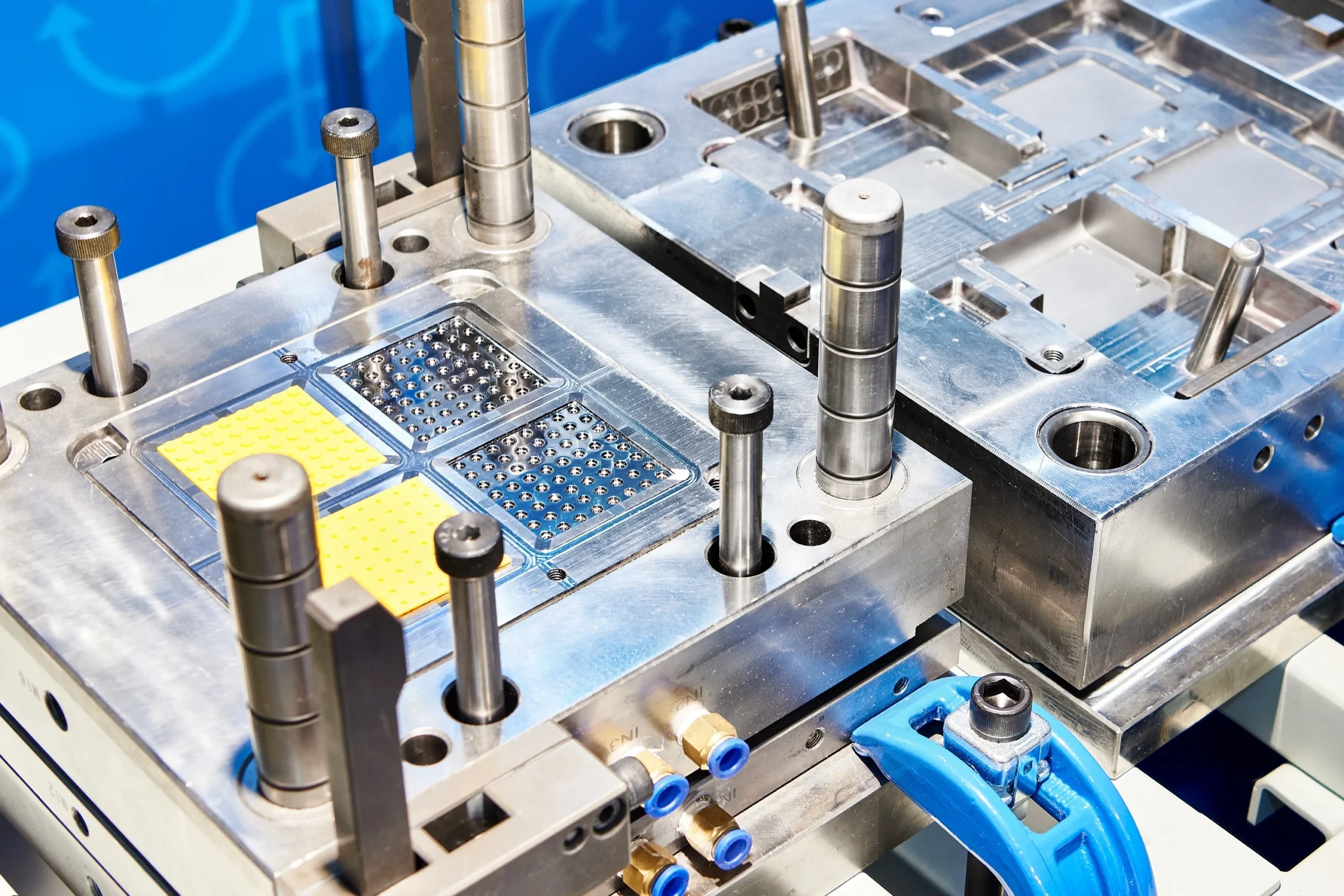 Close-up of a metal injection mold machine used in manufacturing. The mold has complex patterns and openings for shaping materials. It features several cylindrical components and is set against a blue background.