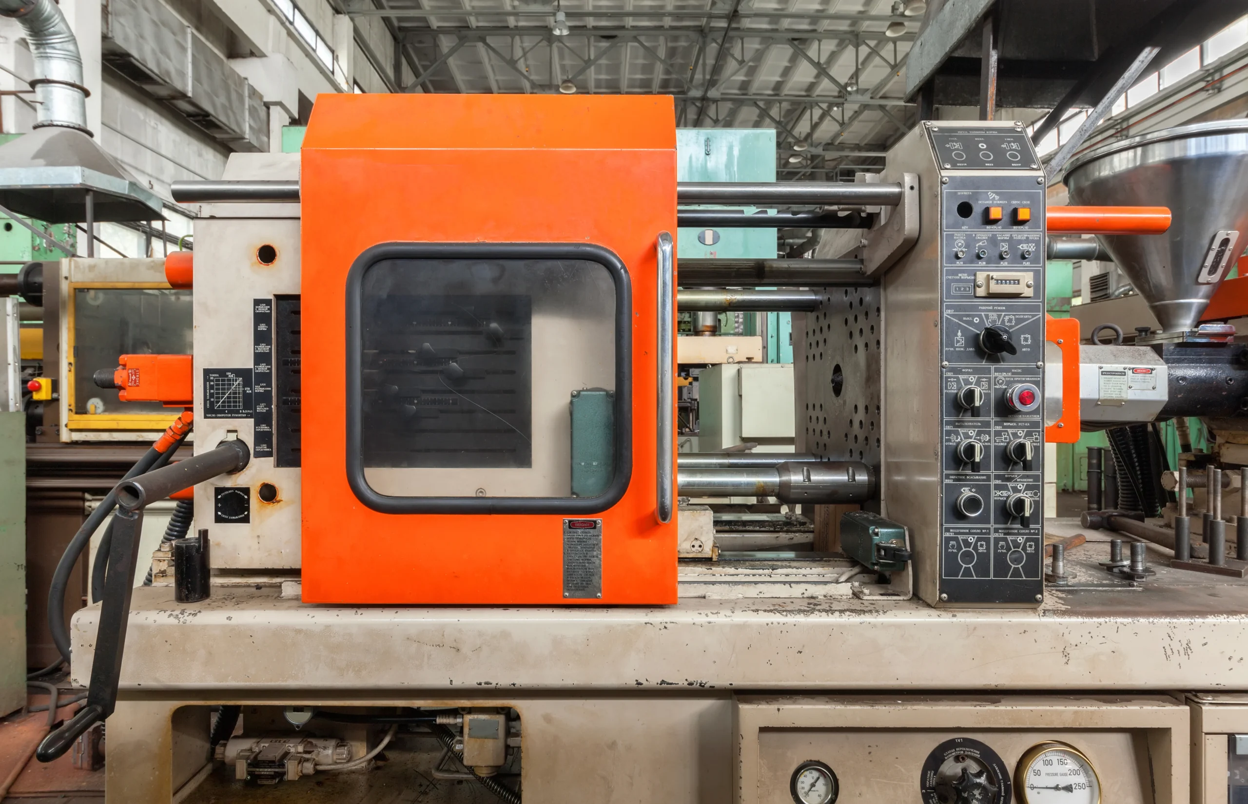 A close-up of an industrial injection molding machine with a prominent orange cover. Control panels with various knobs and switches are visible on the right side, and the factory's ceiling is seen in the background.