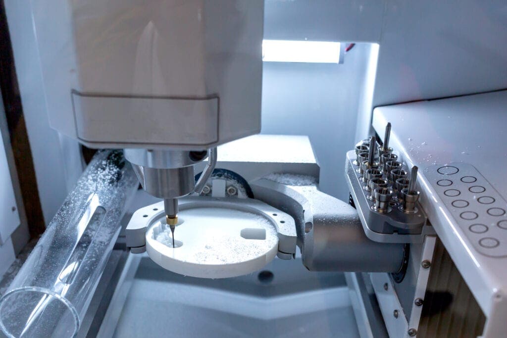 A dental milling machine in operation, carving a white disc. The machine is equipped with various tools positioned on the right. The lighting is bright, illuminating the precise and automated milling process inside the enclosed space.