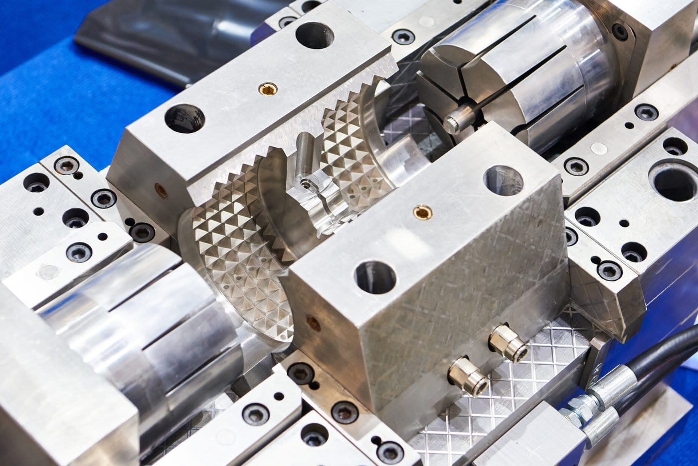 Close-up of a precision metal mold with cylindrical and textured surfaces, used for industrial manufacturing. The mold components are arranged neatly on a blue background, showcasing intricate details and craftsmanship.