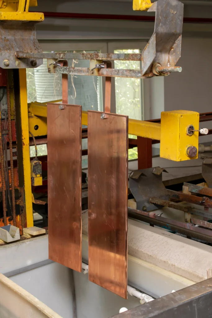 Copper Plates Are Suspended In An Industrial Electroplating Setup, With A Yellow Metal Frame Holding The Plates Above A Liquid-Filled Tank. Machinery And Pipes Are Visible In The Background.