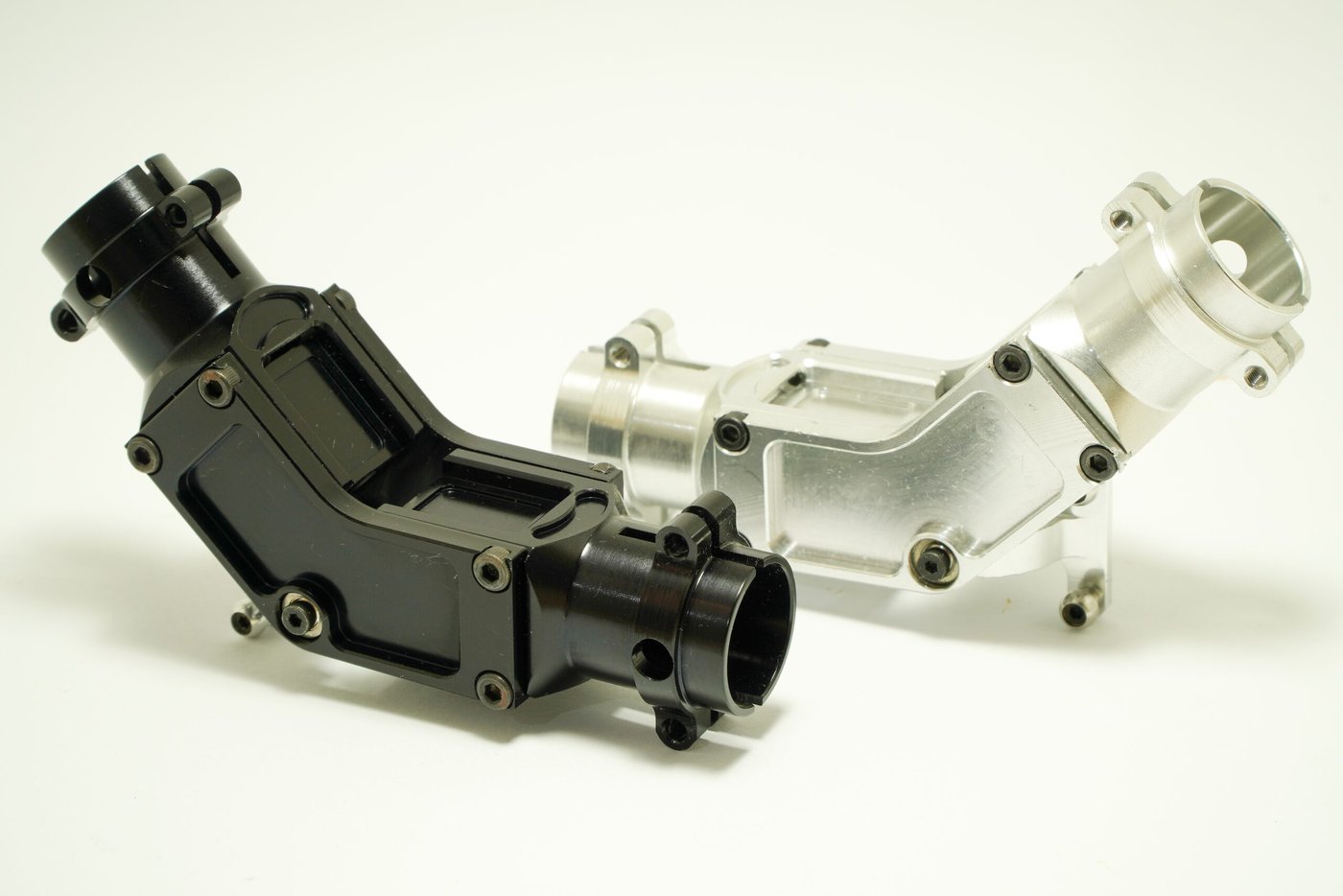 Two metallic, L-shaped engine components are displayed on a light background. One is black and the other is silver, featuring multiple bolt holes and detailed machining, suggesting precision engineering.