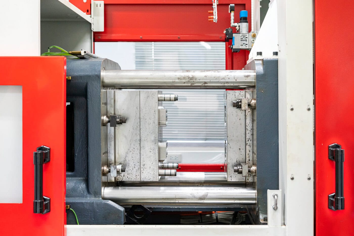 Injection molding machine with metal components and a red and white outer casing. The image shows a mold inside, with visible hydraulic cylinders and various mechanical elements, used for shaping materials in manufacturing processes.