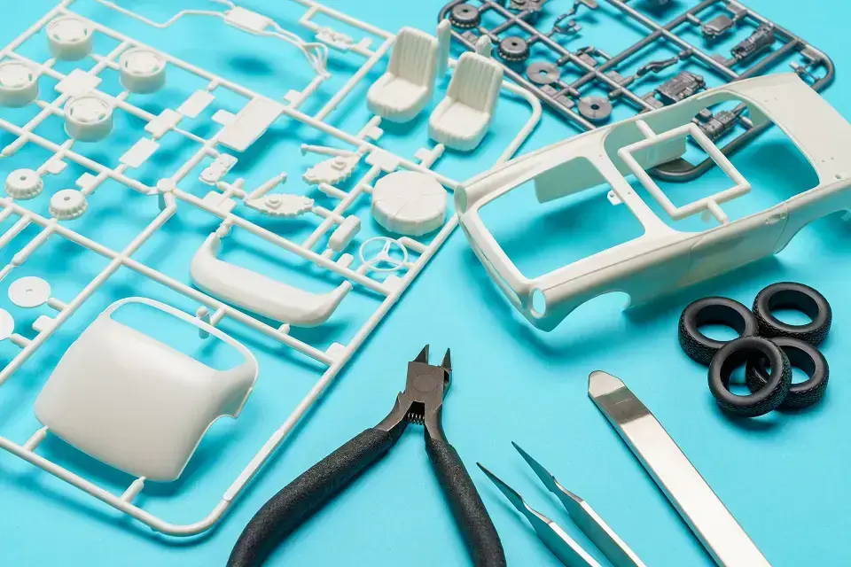 Plastic Parts And Tools For Assembling A Scale Model Car Are Spread Out On A Blue Surface. White Car Components, Black Tires, Clippers, And Tweezers Are Visible.