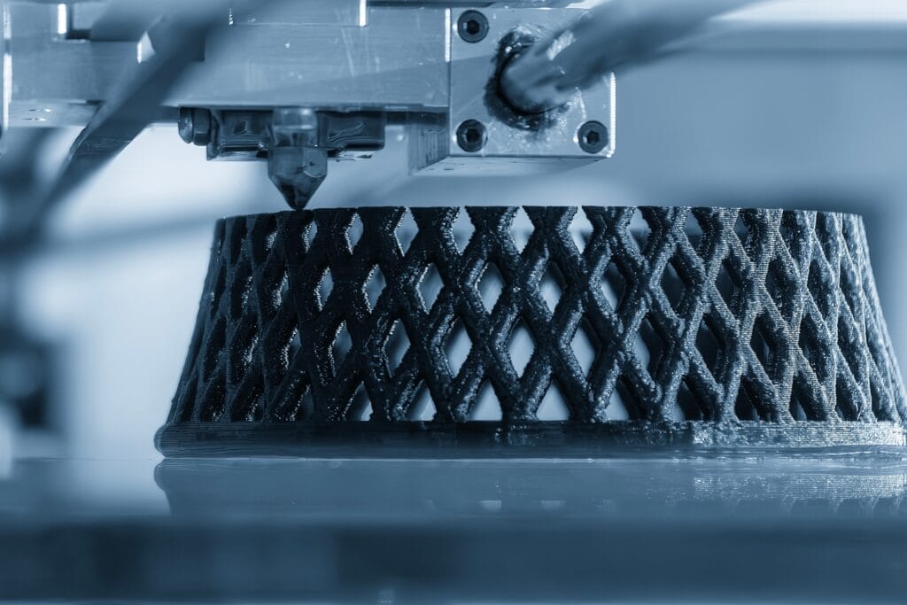 A 3D Printer Creates A Lattice-Patterned Object With A Black Filament. The Focus Is On The Intricate Design Being Printed From The Nozzles. The Setting Appears To Be A Workshop Or Lab, Captured In A Cool Blue Tint.