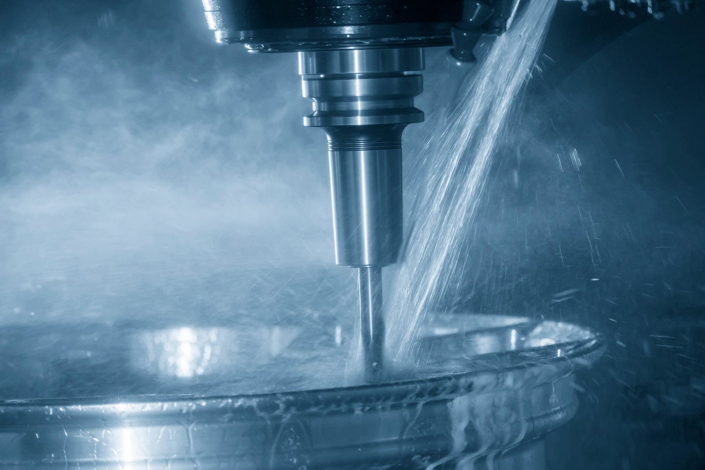 A CNC machine in action, cutting into a metal surface with precision. Water sprays around the machine, creating a misty effect. The scene is lit with a cool, blue-tinted light, highlighting the industrial atmosphere.