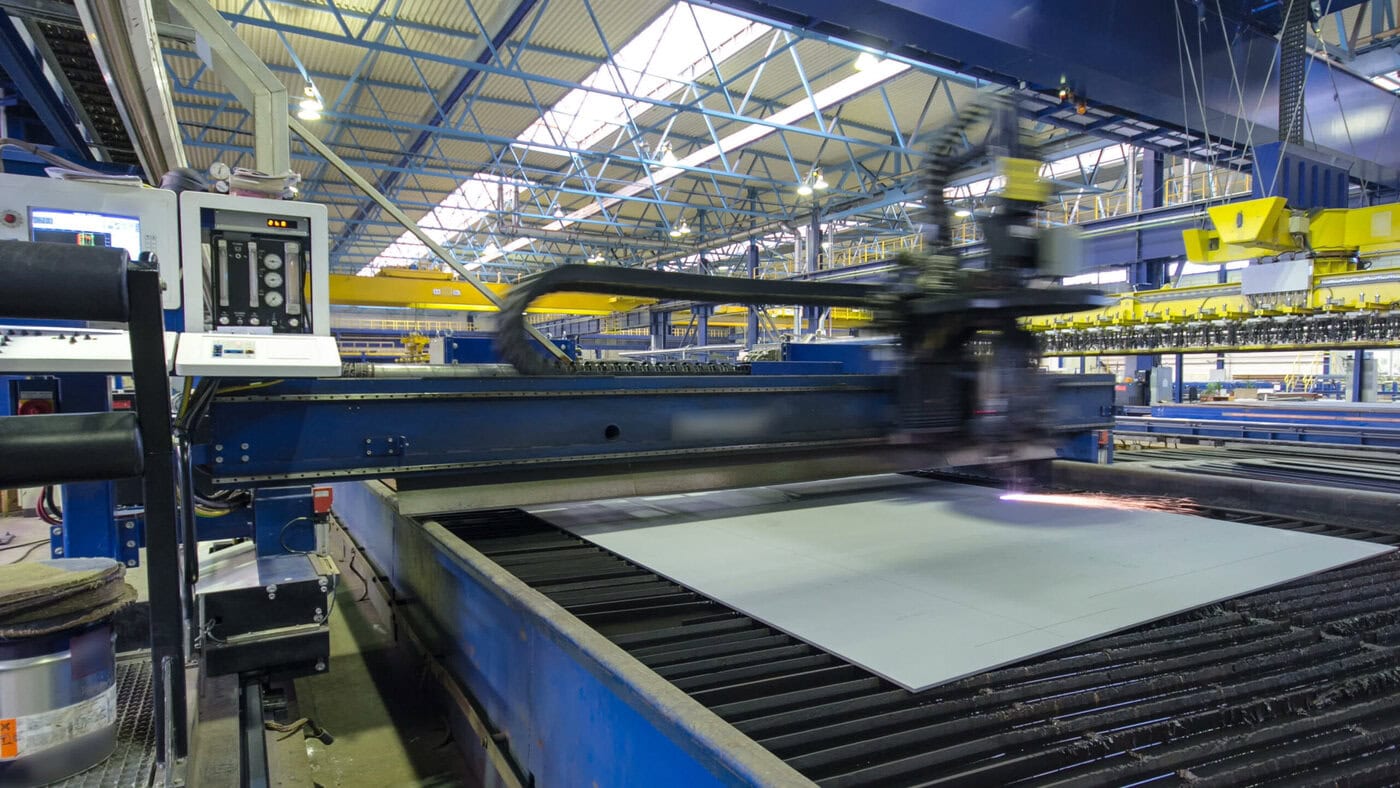 A large industrial machine in a factory is cutting a metal sheet on a production line. The factory is spacious with blue and yellow structural elements and bright overhead lighting.