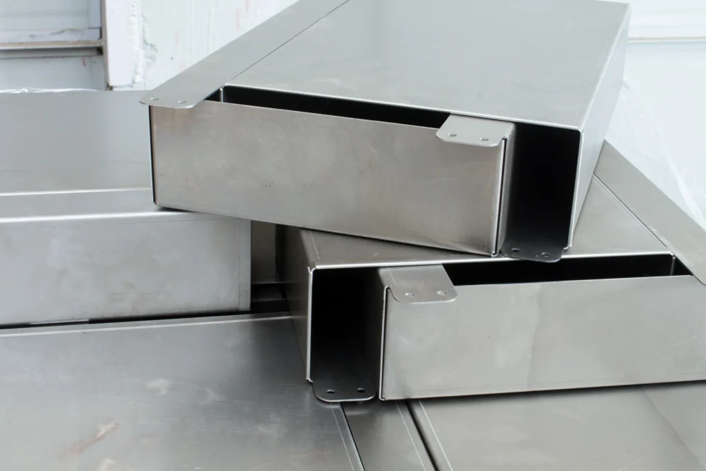 Stacks Of Large Rectangular Metal Ducts Arranged Haphazardly On A Surface. The Ducts Have A Smooth, Shiny Finish And Are Positioned In A Way That Two Are Slightly Open, Displaying Their Hollow Interior.