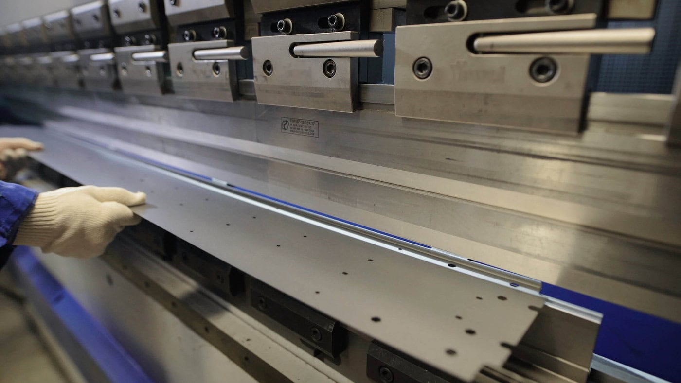 Close-up of a person wearing gloves operating a metal bending machine, aligning a sheet of metal with precision. The machine shows several clamps and tools for the bending process.