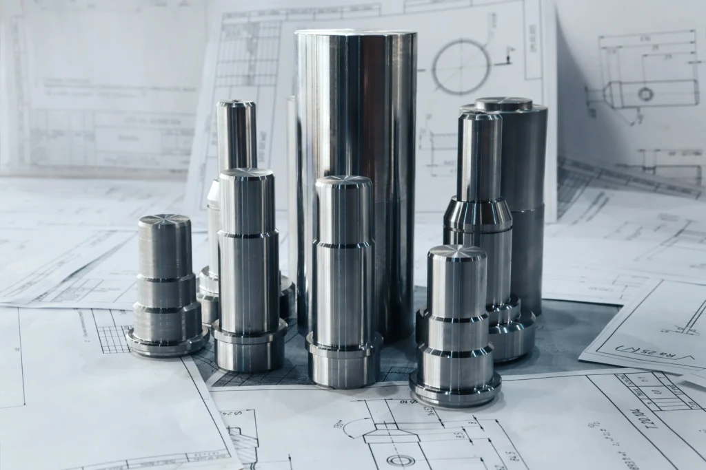 A Group Of Cylindrical Metal Components Of Varying Sizes Placed On Detailed Technical Blueprints. The Metallic Parts Have A Shiny, Polished Finish, Contrasting With The Monochrome Design Sketches Underneath Them.