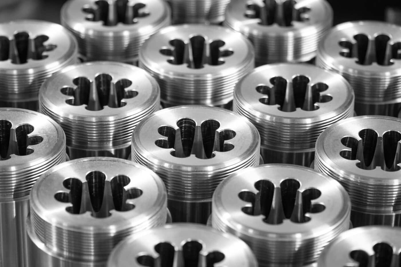 Close-up of multiple metallic threaded cylinders with petal-shaped cutouts on top, arranged in a grid pattern. The shiny surfaces reflect light, highlighting the precision of their design.