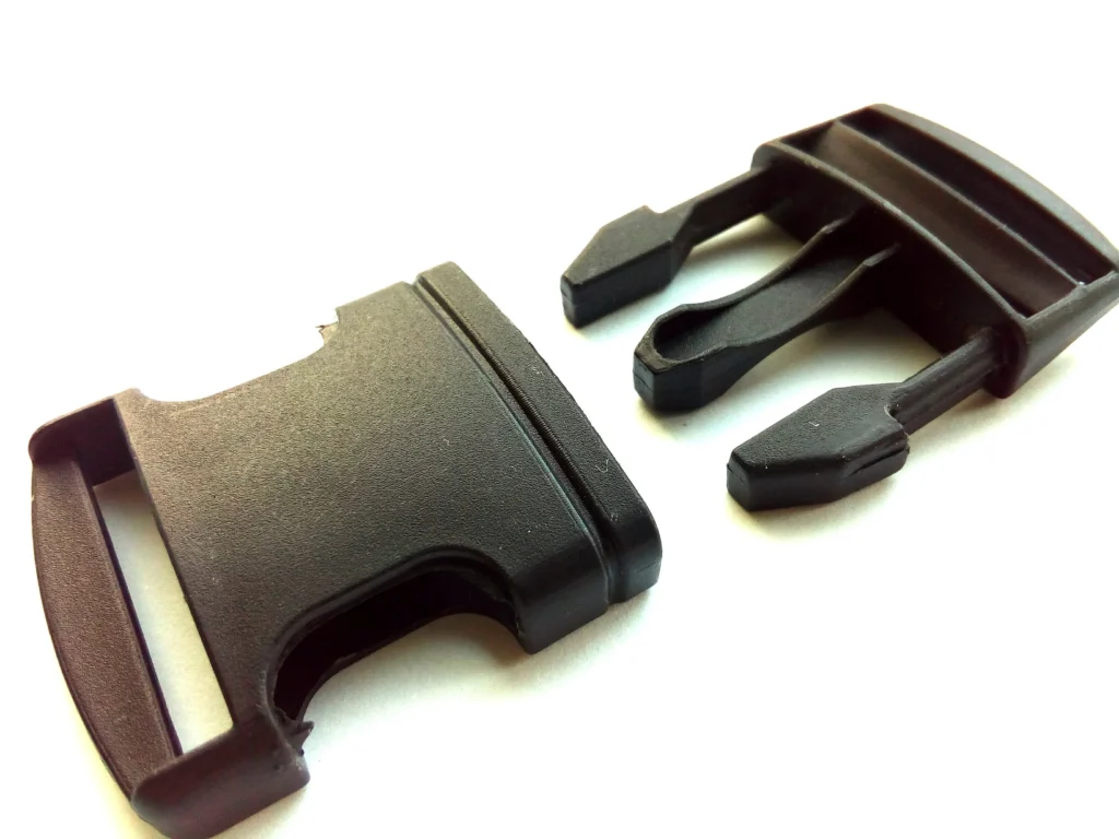 Close-Up Of A Black Plastic Side-Release Buckle, Featuring Two Separate Interlocking Parts Placed On A White Surface. The Buckle Is Commonly Used For Securing Straps On Backpacks And Other Gear.