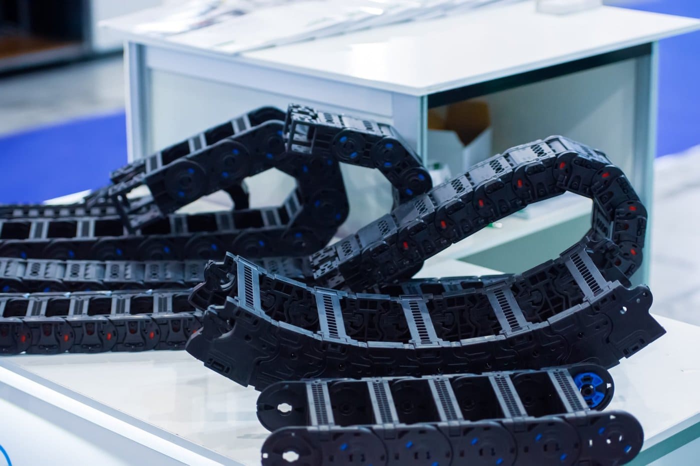 A coiled black plastic cable carrier sits on a white table, with more sections of cable carriers visible in the background. The table is in an indoor setting with blue flooring visible.