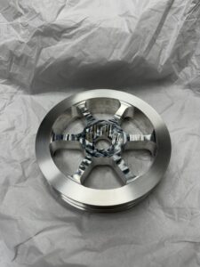 A Polished Metal Wheel With A Five-Spoke Design, Reminiscent Of Battlebots Engineering, Rests On A Crumpled White Paper Background. The Center Showcases A Textured, Multicolored Pattern.